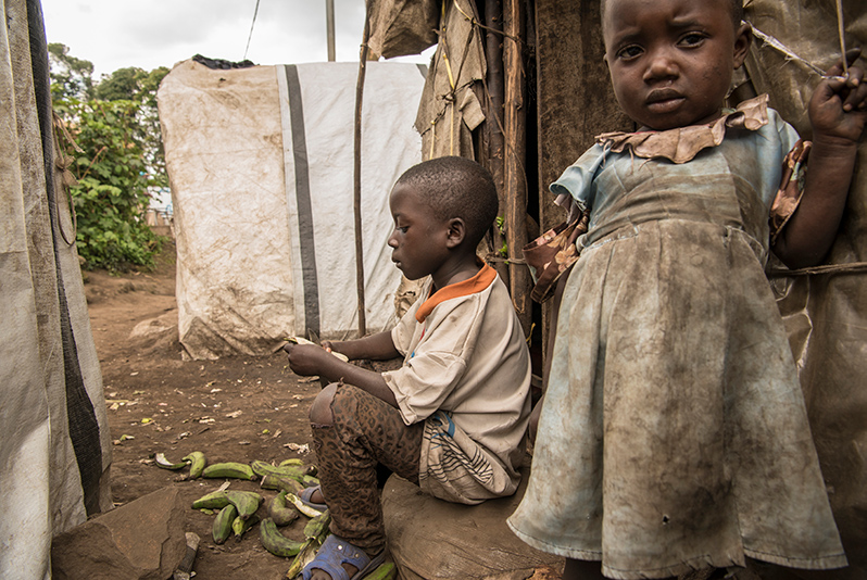Wokowo Mbikameshe skreller kokebananar. Han bur i ein flyktningleir for internt fordrivne kongolesarar. Til høgre står tre år gamle Tatuna Nabwagala.